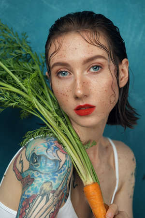 Portrait of tender female fashion model sitting at the blue table with carrot plant. Freckled girl with tattoo directly look at camera in front of the blue background.の写真素材