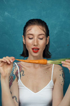 Fashion female freckled model sitting in front of blue background and going to bite carrot.の写真素材