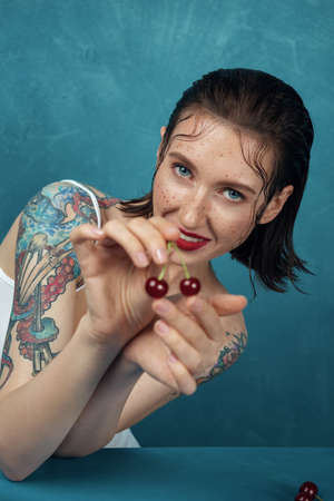 Portrait of young fashion model with cherry berries. Freckled girl is looking directly in camera in front of the blue background.の写真素材