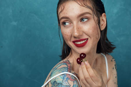 Close-up portrait of young smiling fashion model with cherry berries. Freckled girl is posing in front of the blue background.の写真素材
