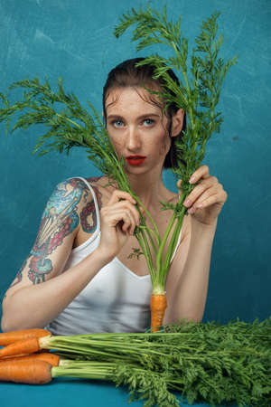 Fashion female tattooed model sitting at the blue table with bunch of carrots. Freckled girl directly look at camera in front of the blue background.の写真素材