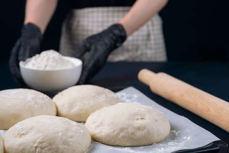 Woman in checkered apron and black gloves holds a flour bowl behind baking tray with pieces of yeast dough. Process of making bakery. The Adjarian Khachapuri Recipe â Georgian cheese bread.の写真素材