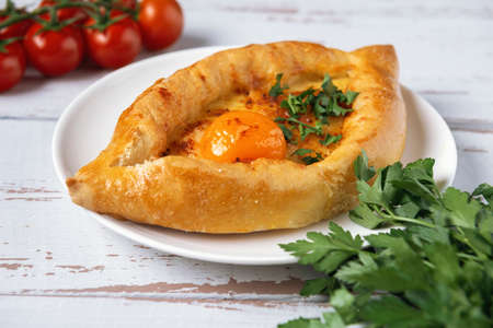 White plate with Adjarian Khachapuri â Georgian cheese bread decorated with tomatoes and parsley on white Provence style background. Baked open pie with cheese and egg yolk filling.の写真素材