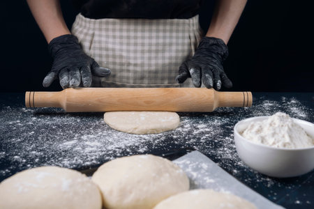 Woman in checkered apron and black gloves rolling out yeast dough with a rolling pin. Process of...