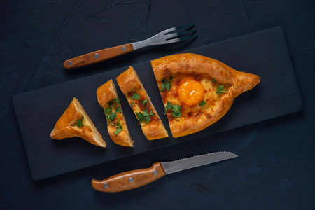 Sliced Adjarian Khachapuri â Georgian cheese bread with knife and fork on dark blue textured background. Baked open pie with cheese and egg yolk filling. Top view. Flat lay.の写真素材