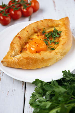 Adjarian Khachapuri â Georgian cheese bread decorated with tomatoes and parsley on white plate and Provence style background. Baked open pie with cheese and egg yolk filling.の写真素材