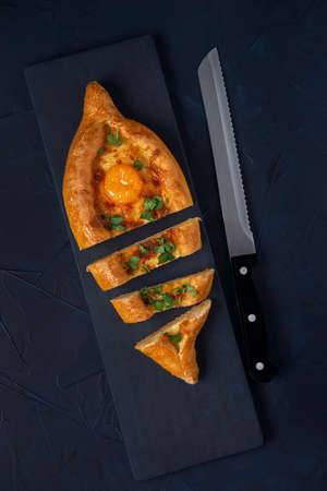 Sliced Adjarian Khachapuri â Georgian cheese bread with kitchen knife on dark blue textured background. Baked open pie with cheese and egg yolk filling. Top view. Flat lay.の写真素材
