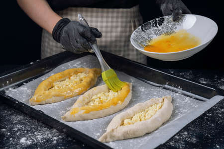 Woman in checkered apron and black gloves covers raw bakery khachapuri with egg yolk. The Adjarian Khachapuri Recipe â Georgian cheese bread.の写真素材