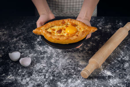 Woman's hands hold black plate with Adjarian Khachapuri â Georgian cheese bread. Baked open pie with cheese and egg yolk filling.の写真素材