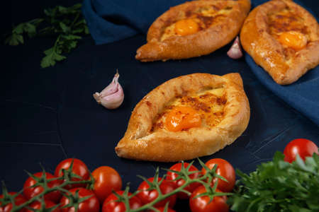 Adjarian Khachapuri â Georgian cheese bread with tomatoes, parsley and garlic on dark blue textured background. Baked open pie with cheese and egg yolk filling.の写真素材