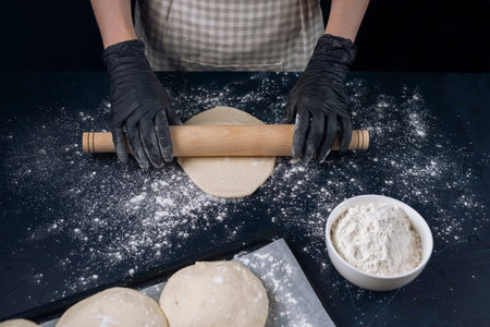 Woman in checkered apron and black gloves rolling out with a rolling pin. Process of making bakery....