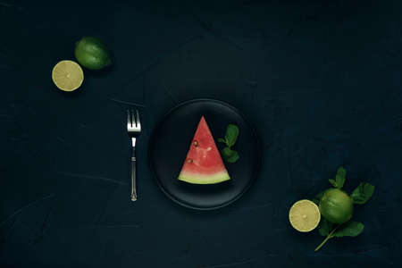 Black plate with slice of watermelon, fork, mint and lime on dark textured background. Flat lay.の写真素材