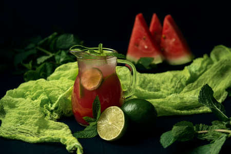 Glass jug with ice-cold watermelon juice, lime, mint, watermelon slices and green gauze on the dark textured backdrop.の写真素材