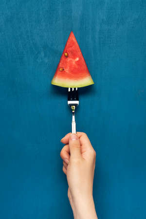 Womanâs hand holds work with watermelon slice on blue wooden background. Flat lay. Top view.の写真素材
