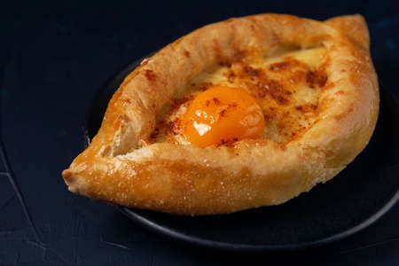 Black plate with Adjarian Khachapuri â Georgian cheese bread on dark blue textured background. Baked open pie with cheese and egg yolk filling.の写真素材
