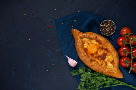 Adjarian Khachapuri â Georgian cheese bread with tomatoes, parsley, garlic and peppercorns on dark blue textured background. Baked open pie with cheese and egg yolk filling. Top view. Flat lay. Space for text.の写真素材