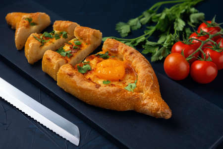Sliced Adjarian Khachapuri â Georgian cheese bread decorated with tomatoes and parsley on dark blue textured background. Baked open pie with cheese and egg yolk filling.の写真素材