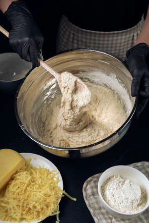 Woman in checkered apron and black gloves mixing yeast dough with wooden spoon. Process of making bakery. The Adjarian Khachapuri Recipe â Georgian cheese bread.の写真素材