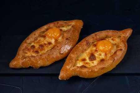 Adjarian Khachapuri â Georgian cheese bread. Two baked open pies with cheese and egg yolk filling. Bakery with wheat flour on dark blue textured background.の写真素材