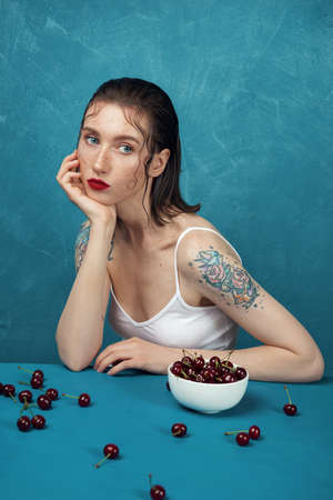Young dreamed woman sitting at the table with cherry berries in front of the blue background.の写真素材