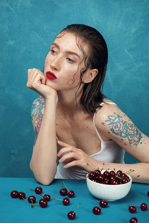 Young tattooed woman in white shirt sitting at the table with cherry berries in front of the blue background.の写真素材