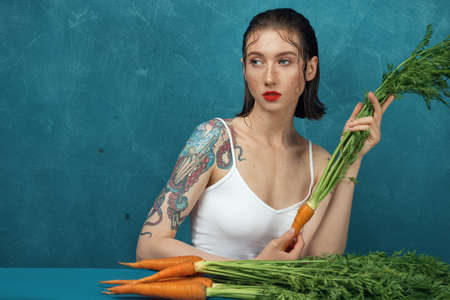 Fashion female tattooed model sitting at the blue table with bunch of carrots.の写真素材