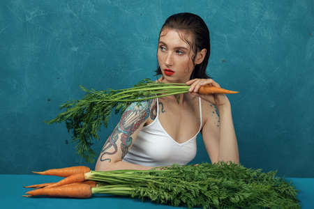 Fashion female tattooed model sitting at the blue table with the bunch of carrots.の写真素材