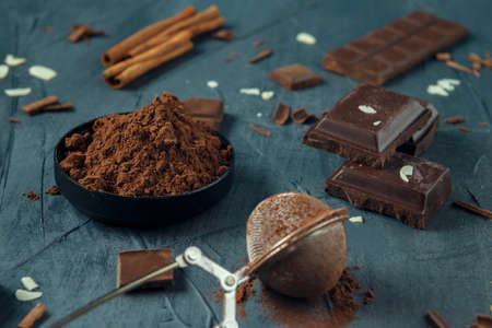 Bowl with cocoa, strainer, pieces of chocolate, chocolate shavings, cinnamon, and almond flakes on the grey textured background.の写真素材