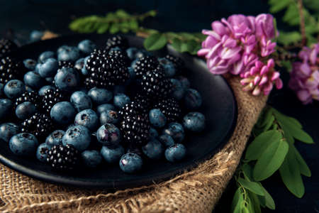 Black plate with blueberry, blackberry and acacia flowers on the burlap napkin and the dark textured background.の写真素材
