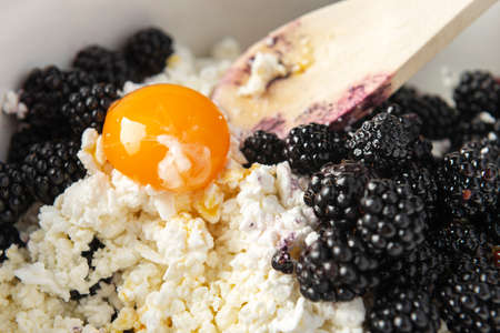 Blackberry, cottage cheese, egg and wooden spoon into the bowl before mixing. Cooking process. Cottage cheese casserole making.の写真素材