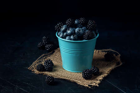 Blue bucket with blueberry and blackberry on burlap napkin and the dark textured background.の写真素材