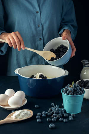 Woman with wooden spoon put blackberry into the bowl with cottage cheese. Cooking process. Cottage cheese casserole making.の写真素材