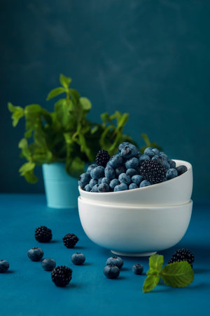 White bowl with mix blueberry and blackberry and blue bucket with mint leaves on the blue background.の写真素材