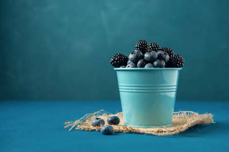 Small bucket with blueberry and blackberry on burlap napkin and blue backdrop.の写真素材
