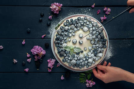 Female hands cut cottage cheese casserole decorated with blackberry, blueberry and acacia flowers on the dark wooden table. Flat lay. Copy space.の写真素材