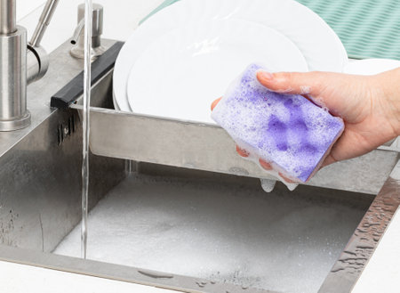 Hand holding a soapy purple sponge over the kitchen sink, ready to wash dishesの写真素材