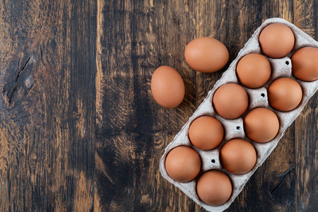 Ten brown eggs sit in a cardboard carton on a rustic wooden table, ready for cooking or bakingの写真素材