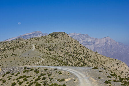 Omani dirt patch street in Oman, trough a beautiful mountains landscape. On the left, the moon, clearly visible even in the day light.の写真素材