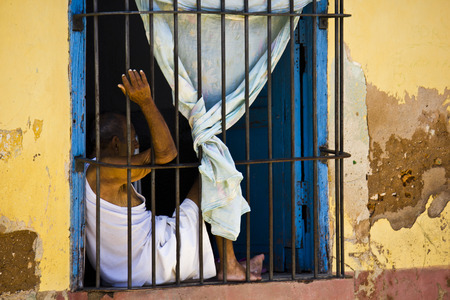 Trinidad, August 18, 2012. Life in trinidad, Cuba. Men behind its window. Daily Life in Trinidad, Cuba. Men inside its house's window.  The sbars make it seemd like a prison.のeditorial素材