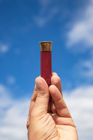 Hand holding a red shotgun shell with blue sky and clouds in the background