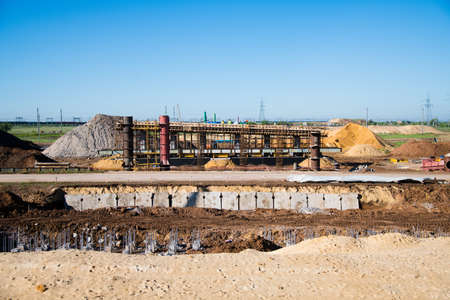 Construction of the interchange of a road bridge across the river. Builders and construction equipment on the construction site. The support of the bridge against the blue sky.の写真素材