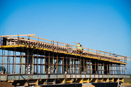 Construction of the interchange of a road bridge across the river. Builders and construction equipment on the construction site. The support of the bridge against the blue sky.の写真素材