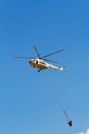 Firefighting helicopter with fire bucket flies against a clear blue skyの写真素材