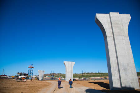 Construction of the interchange of a road bridge across the river. Builders and construction equipment on the construction site. The support of the bridge against the blue sky.の写真素材