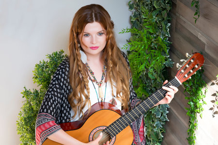A beautiful girl with red curly hair stands and holds a seven-string guitar in her hands. Studio...