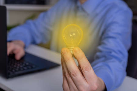 A young man is sitting next to a laptop computer, his hand holding a glass light bulb. Concept idea.の写真素材