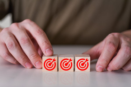 A businessman holds a wooden cube with a goal icon on a wooden table. Goals and planning for success in the marketing business, achieving an objective concept. Close-up and free space for copyingの写真素材