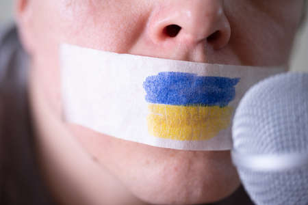 Tape over his mouth with the flag of Ukraine, trying to speak into a microphone. The concept of freedom of the press in Ukraine.の写真素材