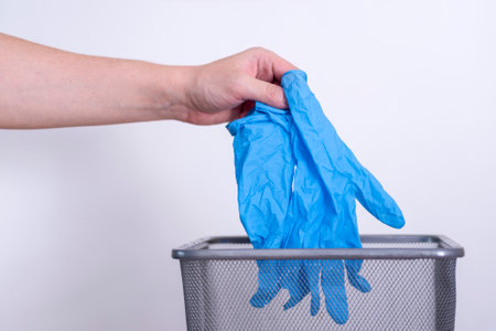 A man's hand throws blue medical gloves into a trash can against a gray background. medical waste.の写真素材