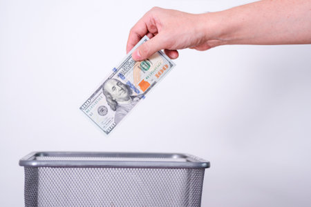 A hand throws dollar bills into a trash can against a gray background. The concept of unnecessary currency.の写真素材
